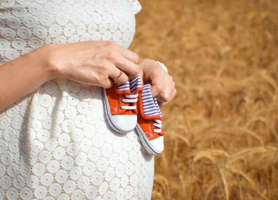 Pregnant woman holding baby shoes up to her belly in a field. If you’ve been experiencing postpartum anxiety in Charlotte, NC, meet with a compassionate postpartum therapist in North Carolina, South Carolina, or Florida! You deserve this. 28207 | 28211 | 28036