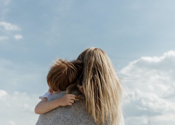 Mom with back turned to camera and long blonde hair, hold her son in her arms while he rests his head on her should. The sun is out and there are clouds in the sky. Mom. Mother's love. children.