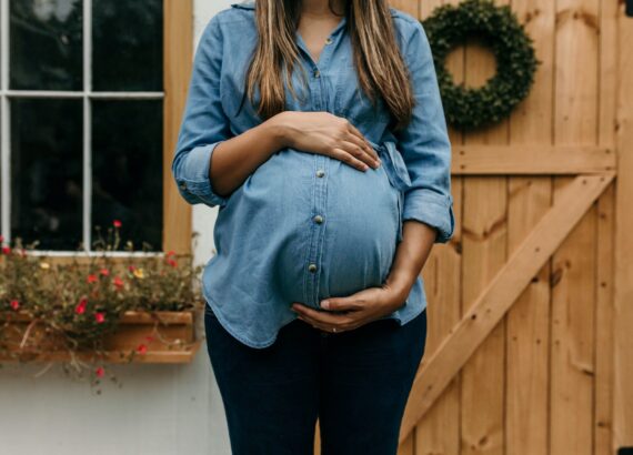 Pregnant woman standing outside in front of a white house with a window and a light wooden gate. New mom anxiety. Postpartum counseling near Cary, North Carolina.