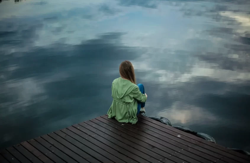 A woman sitting on a dock, gazing at the water, reflecting on the complexities of her traumatic experiences.
